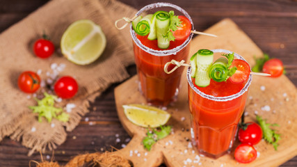 Two glasses of fresh organic tomato juice decorated with raw tomatoes, cucumber and leaves on a rustic wooden cutting board