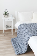 Fototapeta na wymiar Bed with white linen and grey knitted woolen merino chunky blanket. Light stylish cozy scandinavian bedroom interior. Blanket of thick yarn. Geometric glass florarium with plants on bedside table.