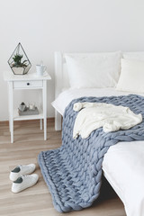 Bed with white linen and grey knitted woolen merino chunky blanket. Light stylish cozy scandinavian bedroom interior. Blanket of thick yarn. White knitted sweater and sneakers on the bed.