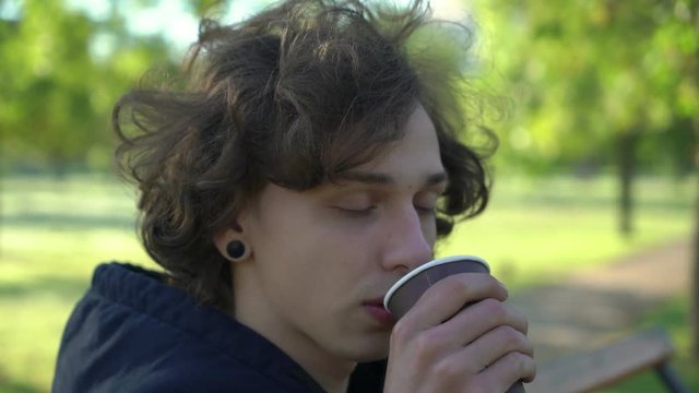young boy sitting on a banch and drink coffee in the morning in the park, 4k