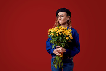 Cheerful young lady in blue clothes, black beret and round glasses being excited to get bouquet of yellow roses on women's day or birthday isolated over red background. French style girl 