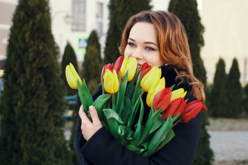 Fototapeta premium A young girl in a black coat with red hair is holding a bouquet of yellow and red tulips and smiling. Beautiful girl in a black coat with a bouquet stands on the background of green fir trees.