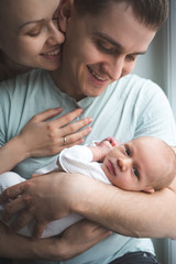 Obraz na płótnie Canvas Man holding on hands a newborn. Woman hugs a man from behind. Sitting next to the window. Mom, dad and baby. Portrait of young family. Happy family life. Man was born.