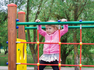 Fototapeta premium A little girl learns to climb ladders and crossbars on the playground of the local park.