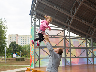 Fototapeta premium Father throws up his Little girl baby by his hands. Dad is a great good interesting attraction for his little daughter. The child smiles in flight.