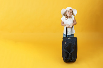 Little child girl blonde is ready for travel and stands with a suitcase. Yellow background