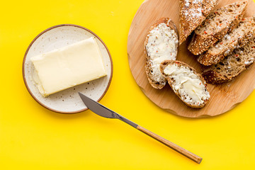 Wholegrain bread on cutting board with butter on plate, slice of bread with butter on yellow background top view