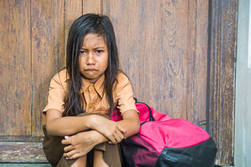 7 or 8 years child in school uniform sitting outdoors on the floor crying sad and depressed holding her backpack suffering bullying and abuse problem feeling helpless