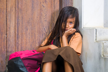 7 or 8 years child in school uniform sitting outdoors sad and depressed with her backpack on the stairs suffering bullying and abuse problem feeling alone and helpless