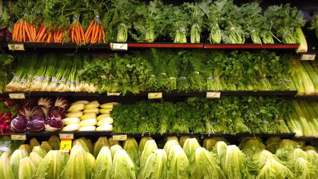 Dolly shot of supermarket vegetable display. Green grocery fresh for sale at local market. Shelves of various organic greens. Lettuce, celery and carrots displayed.
