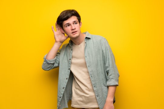 Teenager man over yellow wall listening to something by putting hand on the ear