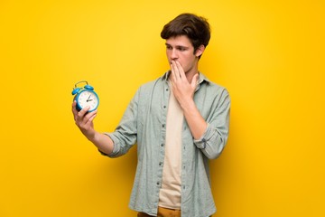 Teenager man over yellow wall holding vintage alarm clock