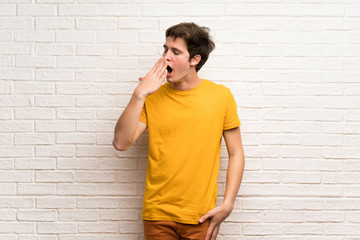 Teenager man over white brick wall yawning and covering wide open mouth with hand