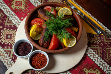 Chicken cutlet with vegetables in a clay pot on a light wooden tray, on a wooden table with a tablecloth with national patterns.