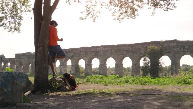 Cyclist with bike standing on a rock under a tree reading a map in front of ancient roman aqueduct. Young attractive athletic man with orange sportswear and backpack in parco degli acquedotti in Rome