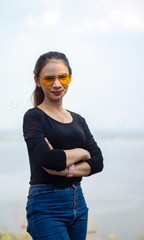 Women white skin lovely brown hair a small woman red lips wearing a black shirt wearing a blue gene woman Wearing yellow glasses stand  poses photography portrait which has water in back in the park.