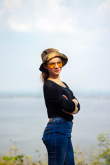 Women white skin lovely brown hair red lips black shirt wearing a blue gene woman helmeted wearing yellow glasses stand  poses photography portrait which has water in back in the park.
