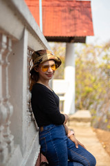 Women white skin lovely brown hair red lips black shirt wearing a blue gene woman helmeted wearing yellow glasses stand  poses photography portrait which has water in back in the park.