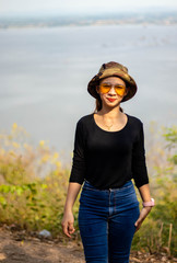 Women white skin lovely brown hair red lips black shirt wearing a blue gene woman helmeted wearing yellow glasses stand  poses photography portrait which has water in back in the park.