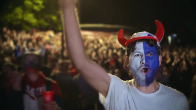 Football fan shouts, rejoices goal favorite 4K team. Person paint face jump in delight victory match. Closeup guy screaming furiously, jumping with friend against backdrop crowd winning soccer game 4k