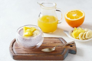 Chia pudding with mango pulp  in beautiful glasses with green mint leaves and cut fresh ripe tropical fruit, orange juice for breakfast on a light background - raw vegetarian sweet organic dessert. 