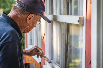 Active old man is concentrating for painting window frame on wooden house