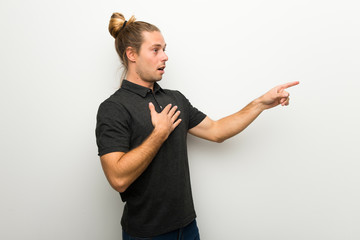 Blond man with long hair over white wall pointing finger to the side with a surprised face