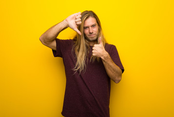 Blond man with long hair over yellow wall making good-bad sign. Undecided between yes or not