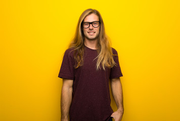 Blond man with long hair over yellow wall with glasses and happy