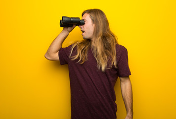 Blond man with long hair over yellow wall and looking in the distance with binoculars