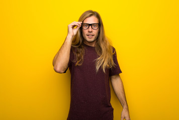 Fototapeta premium Blond man with long hair over yellow wall with glasses and surprised