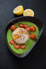 Fototapeta na wymiar Bowl with baked fish medallions and cherry tomatoes on a bedding of green peas puree, studio shot over black stone background