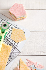 Fabric pentagon on craft mat next stack of pentagon pieces of fabrics, craft tools on white wooden surface, top view.