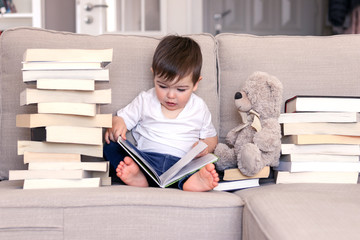 Cute clever little baby boy keen about reading book sitting on sofa with teddy bear toy and piles...