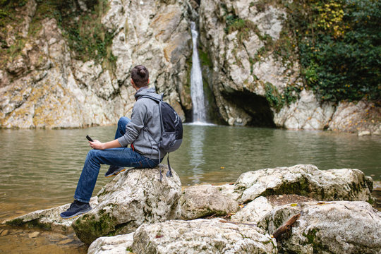 Lonely travaler guy with backpack sits in picturesque place on stone by mountain river and communicates in social networks and smartphone. Concept of unity of modern technology and wildlife