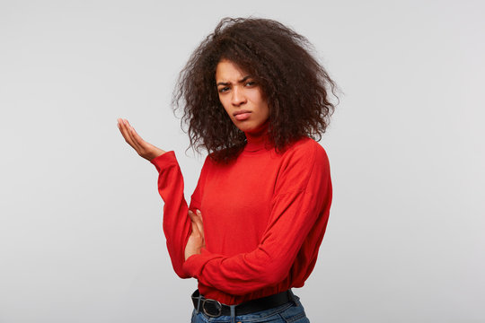 Young dark-skined woman with afro hairstyle in a red longsleeve standing half turned with palm raised up in perplexity with bewilderment puzzeled isolated on a white background