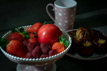 Raspberries, strawberries and pomegranates on a stand with Canele cakes in the background.