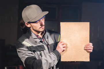 Portrait of a young carpenter in overalls and goggles with a mock up board holding a blank sign in...