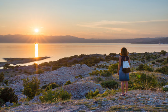 Young woman wearing backpack is enjoying the panoramic view of the sea with sun setting behind the hills. Sunset on the Adriatic sea on island Krk in Croatia. Travel and freedom concept.