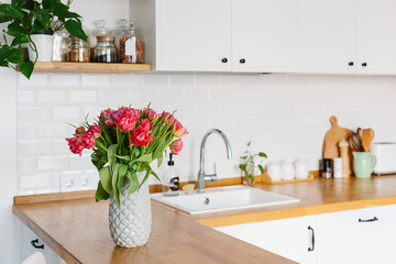Tulips bouquet in vase standing on wooden countertop in the kitchen. Modern white u-shaped kitchen in scandinavian style.