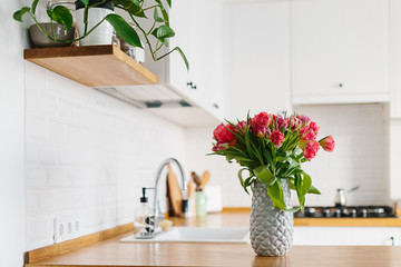 Tulips bouquet in vase standing on wooden countertop in the kitchen. Modern white u-shaped kitchen in scandinavian style.