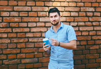 Portrait of a dissatisfied or surprised guy, holding a coffee in his hand against a brick wall, wearing a blue shirt, urban style