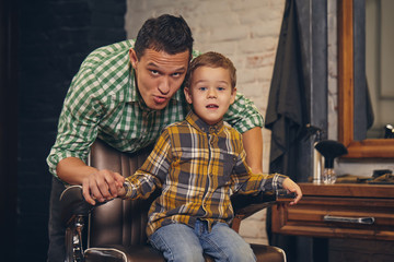 stylish little kid sitting on chair at barbershop with his young father on background