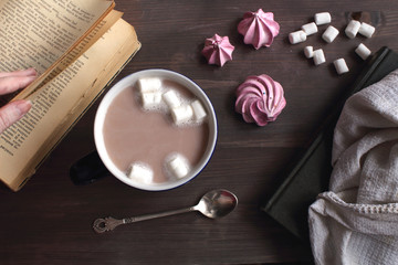 Сocoa coffe with marshmallows in a dark blue cup on a wooden table next to a book, a spoon and meringue top view atmospheric cozy flat lay
