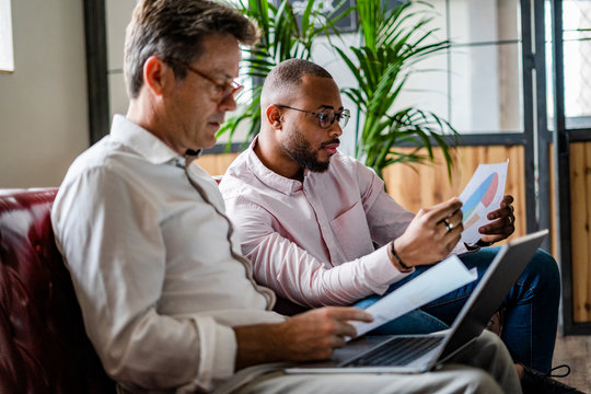Two businessmen using laptop and discussing documents on sofa in loft office