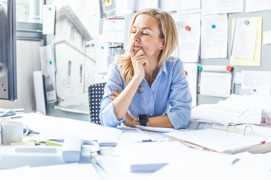 Pensive woman sitting at desk in office surrounded by paperwork