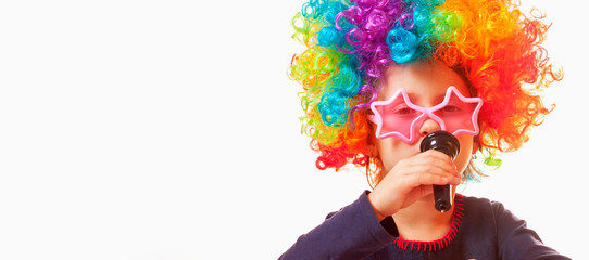 Close up little cute child girl wearing sunglasses with colorful wig singing in microphone. Singer talent and vocal training concept.