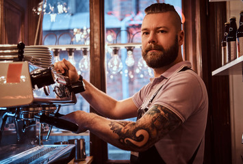 Handsome tattooed barista with stylish beard and hairstyle working on a coffee machine in a coffee...