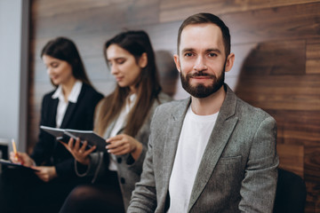 Diverse young businesspeople students friends sitting together in chairs in queue using mobile phones wait job interview or examination. Male showing funny video people laughing, positive emotions