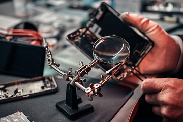 Repairman uses magnifier and tweezers to repair damaged smartphone. Close-up photo of a disassembled smartphone.